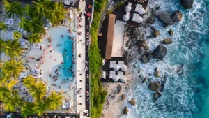 Stunning aerial view of a beachfront pool club in Uluwatu surrounded by palm trees and crashing waves a top-rated destination among the Best Things to Do in Uluwatu, perfect for relaxing after a peaceful Bingin silver class.